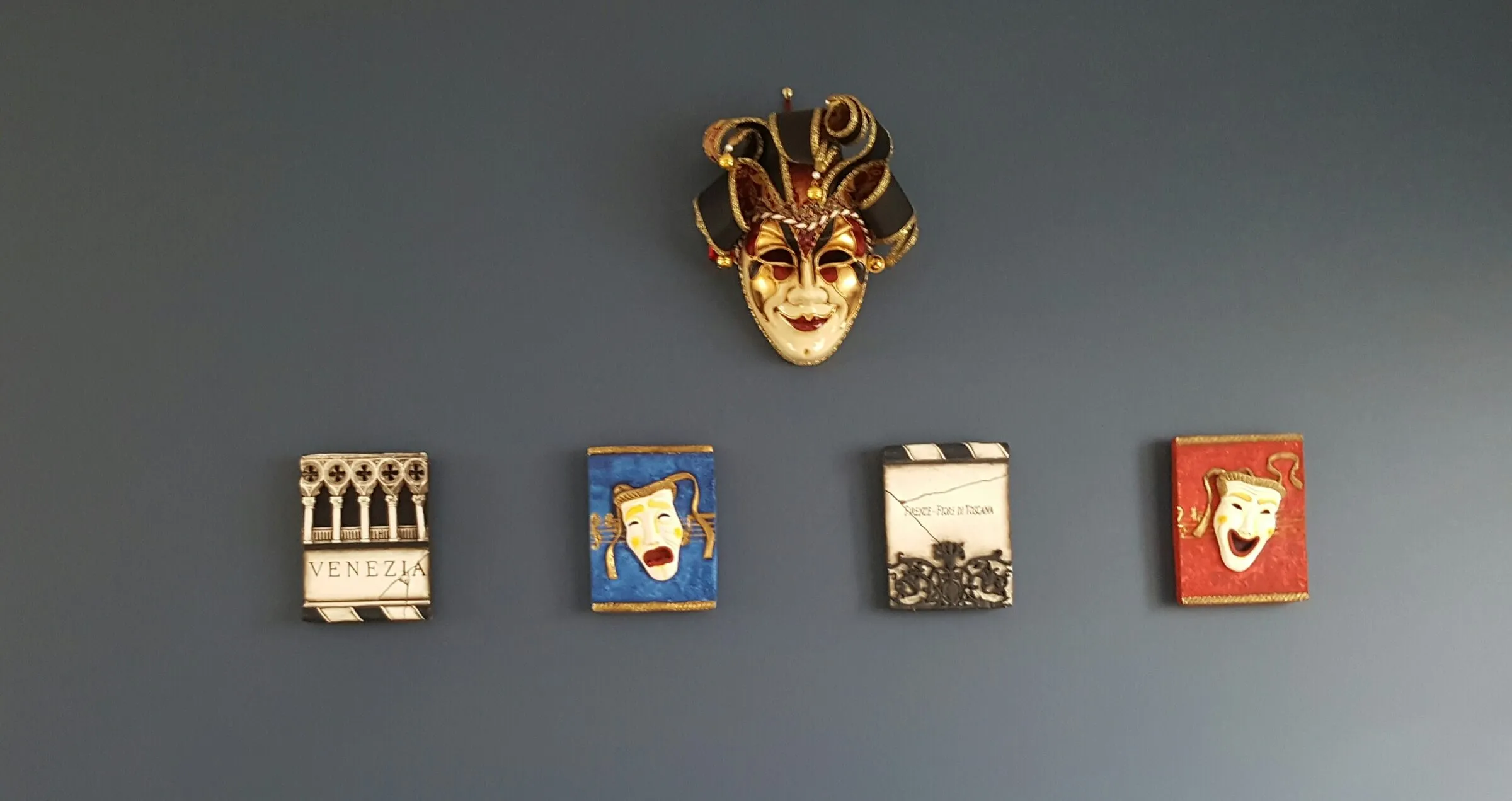 A Venetian mask and five framed pieces hung and levelled across a grey wall.